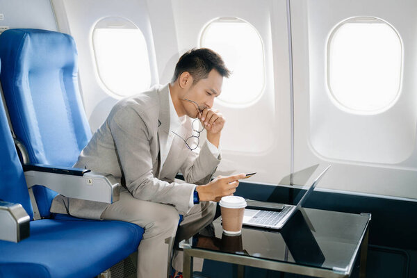 Азиатский бизнесмен сидит в кресле самолета и работает на ноутбуке и с помощью смартфона и кредитной карты для оплаты онлайн собирается в поездку отпуск. 
