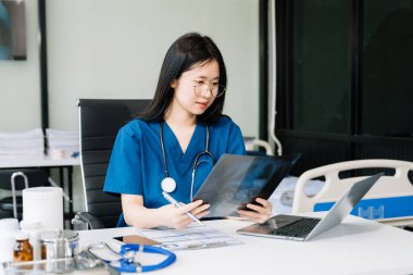 Kendine güvenen genç Asyalı kadın doktor masada oturmuş dizüstü bilgisayar ve röntgenle çalışıyor..