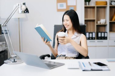 Genç çekici Asyalı kadın ofis çalışanı modern ofiste çalışıyor ve kahve içerken kitap okuyor.