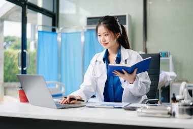 Kendine güvenen genç Asyalı doktor masada oturuyor dizüstü bilgisayar ve not defteriyle çalışıyor.