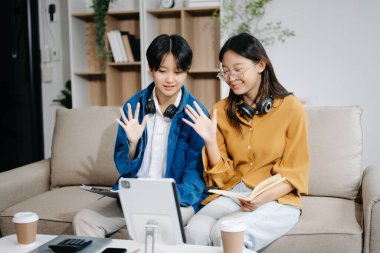 Üniversite kütüphanesinde okuyan iki genç Asyalı öğrenci, masada oturuyorlar, tablet bilgisayar kullanıyorlar, kanepede görüntülü sohbet ediyorlar.