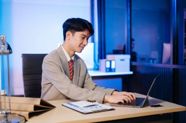 Asian Businessman analyzing reports in a modern office, working on a laptop computer. Perfect for corporate, business, and over time concepts