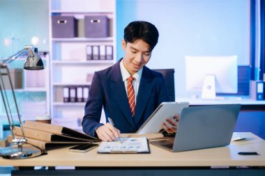 Asyalı İşadamı, modern bir ofiste tabletli bir dizüstü bilgisayar üzerinde çalışan raporları analiz ediyor. Şirket, iş ve zamanla ilgili kavramlar için mükemmel.