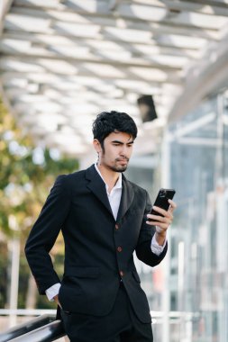 Takım elbiseli genç işadamı, modern bir şehrin dışında duruyor, akıllı telefon kullanıyor.