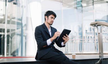 Takım elbiseli genç bir işadamı, dışarıda modern bir şehirde oturuyor, tablet bilgisayar kullanıyor. 