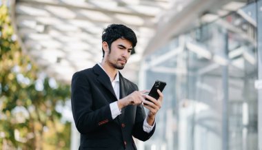 Takım elbiseli genç işadamı, modern bir şehrin dışında duruyor, akıllı telefon kullanıyor.