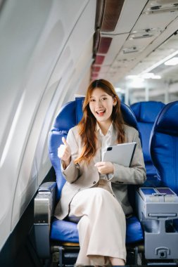Genç Asyalı iş kadını dijital tabletle uçakta baş parmağını gösteriyor. 