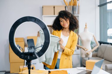 Moda blogcusu konsepti, Afro-Amerikan genç bir kadının evde akıllı telefon ve yüzük lambasıyla video yayınında kıyafet satması. 