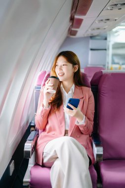 Genç Asyalı kadın yolcu rahat koltukta oturmuş akıllı telefon kullanıyor ve kahve içiyor. 