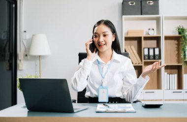 Female entrepreneur using laptop computer and talking on smartphone while working in office  clipart