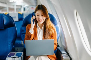 Genç Asyalı iş kadını uçakta dizüstü bilgisayar kullanıyor ve telefonla konuşuyor. 