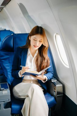 Resmi takım elbiseli Asyalı kadın yolcu rahat koltukta oturup notlar yazıyor. 