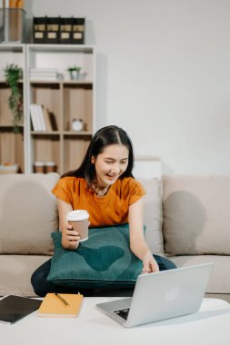 Kanepede oturan Asyalı kadın dizüstü bilgisayar kullanıyor ve kahve içiyor.