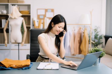 Özel dikim atölyesinde çalışan Asyalı terzi akıllı telefonla konuşuyor ve dizüstü bilgisayar kullanıyor. 
