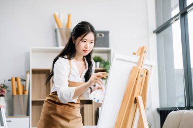 Asyalı kadın ressam sanat atölyesinde resim yapıyor, resim malzemeleri, yağlı boya tabloları, tuval sehpası, resim stüdyosunda veya oturma odasında fırçalı yaratıcı bir alan.