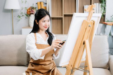 Asyalı kadın ressam sanat atölyesinde resim yapıyor, resim malzemeleri, yağlı boya tabloları, tuval sehpası, resim stüdyosunda veya oturma odasında fırçalı yaratıcı bir alan.
