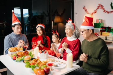 Family celebrating Christmas in a cozy, praying before christmas dinner with festive setting at home with santa hats, gifts, and delicious food clipart