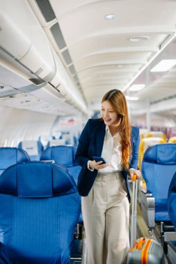 Çekici Asyalı kadın yolcu bavuluyla uçakta duruyor ve akıllı telefon kullanıyor. 
