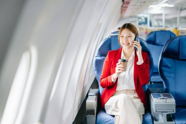 Başarılı bir Asyalı iş kadınının portresi resmi bir takım elbiseyle birinci sınıfta oturmuş akıllı telefon kullanıyor ve kahve içiyor. 