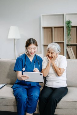 Hemşire ve yaşlı kadın hasta evde dijital tablet kullanıyorlar. Kanepede oturan yaşlı kadınla ilgilenen bir terapist. Tıbbi servis kavramı