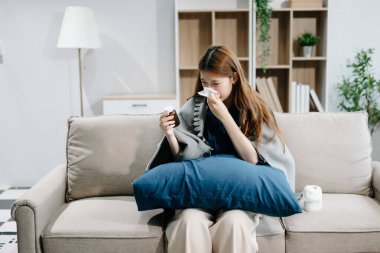 Kanepede battaniyeyle kaplanmış ve mendille hapşıran, grip belirtileri gösteren genç bir Asyalı kadın. Hastalık, sağlık ve iyileşme kavramı