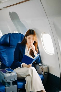 Başarılı bir Asyalı iş kadınının resmi bir takım elbiseyle birinci sınıfta oturmuş kitap okuduğu portresi. 