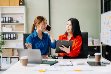 Asyalı iş arkadaşları dizüstü bilgisayar kullanıyor ve akıllı telefondan konuşuyor. Modern ofiste çalışan finansal pazarlama ekibi. 