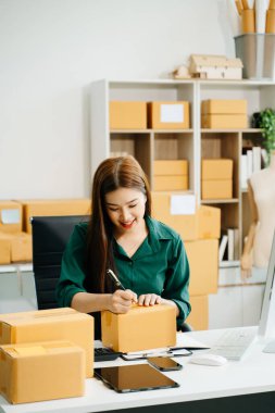 Genç işletme sahibi kadın paket kutuları hazırlıyor ve bilgisayardaki müşterilere teslimat için çevrimiçi siparişleri kontrol ediyor. Çevrimiçi Alışveriş kavramı.