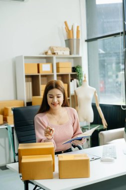 Genç işyeri sahibi kadın, paket kutuları hazırlıyor ve bilgisayardaki müşterilere teslimat için online siparişleri kontrol ediyor. Not defterine not alıyor. Çevrimiçi Alışveriş kavramı