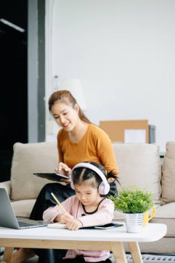 Neşeli anne ve kız kanepede kaliteli zaman geçiriyorlar, gülümsüyorlar, dijital tablet kullanan kadınlar ve deftere yazı yazan kızlar. Aile teması için mükemmel.