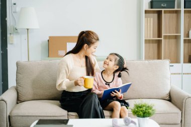Neşeli anne ve kız kanepede güzel vakit geçiriyorlar, gülümsüyorlar ve birlikte kitap okuyorlar. Aile teması için mükemmel.