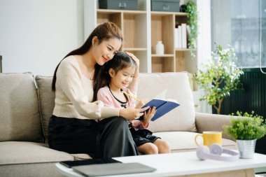 Neşeli anne ve kız kanepede güzel vakit geçiriyorlar, gülümsüyorlar ve birlikte kitap okuyorlar. Aile teması için mükemmel.