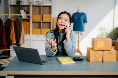 Çevrimiçi satışları yöneten genç bir iş kadını ve modern bir ev ofisinden gelen nakliyat, dizüstü bilgisayar kullanarak ve akıllı telefondan konuşarak 