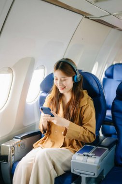 Birinci sınıf uçak koltuğunda birinci sınıf kulaklıklı Asyalı bir kadın akıllı telefon kullanıyor, lüks, rahat ve şık bir seyahat sergiliyor. Havacılık, turizm ve iş hayatı teması için mükemmel.