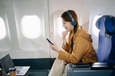 Birinci sınıf uçak koltuğunda birinci sınıf kulaklıklı Asyalı bir kadın akıllı telefon kullanıyor, lüks, rahat ve şık bir seyahat sergiliyor. Havacılık, turizm ve iş hayatı teması için mükemmel.