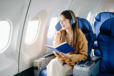 Birinci sınıf uçak koltuğunda kulaklıklı Asyalı bir kadın kitap okuyor, lüks, rahat ve zarif bir seyahat sergiliyor. Havacılık, turizm ve iş hayatı teması için mükemmel.