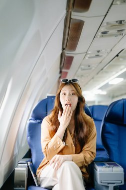 Birinci sınıf uçak koltuğunda oturan Asyalı bir kadın. Şaşırmış bir yüzü var. Lüks, konfor ve zarafet sergiliyor. Havacılık, turizm ve iş hayatı teması için mükemmel.