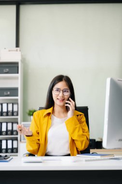 Bilgisayarda çalışan Asyalı genç iş kadını ve ofiste akıllı telefonla konuşan.