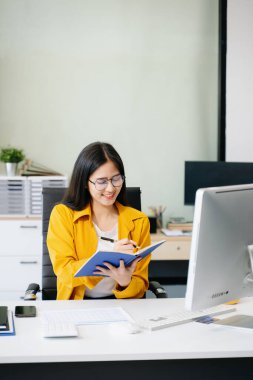 Genç Asyalı iş kadını not defterine not alıyor ve ofiste bilgisayarla çalışıyor.