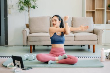 Genç bir Asyalı kadın yoga yapıyor ve paspas ve jimnastik malzemeleriyle esniyor, sağlığını, motivasyonunu ve modern ev ortamında sağlıklı ve aktif bir yaşam tarzını gösteriyor..