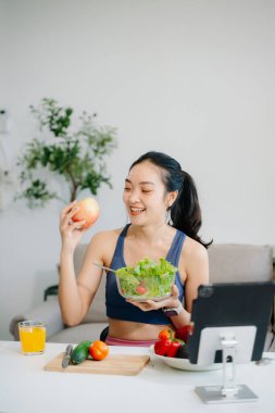 Formda bir Asyalı kadın dijital tabletle çevrimiçi yayın yaparken taze salata, meyve ve meyve suyu içiyor. Parlak, modern bir ev ortamında sağlığın, beslenmenin ve canlılığın vücut bulmuş hali. Sağlıklı bir yaşam tarzı!