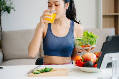 Formda bir Asyalı kadın dijital tabletle çevrimiçi yayın yaparken taze salata, meyve ve meyve suyu içiyor. Parlak, modern bir ev ortamında sağlığın, beslenmenin ve canlılığın vücut bulmuş hali. Sağlıklı bir yaşam tarzı!