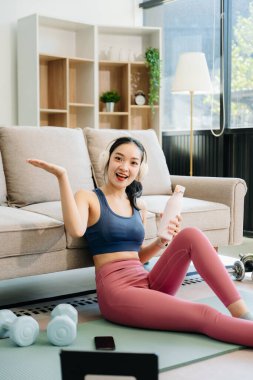 Genç Asyalı bir kadın yoga yaptıktan sonra dinleniyor, su içiyor ve paspas ve jimnastik malzemeleriyle esniyor, sağlığını, motivasyonunu ve modern bir ev ortamında sağlıklı ve aktif bir yaşam tarzını gösteriyor.. 