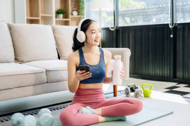 Genç bir Asyalı kadın yoga yaptıktan sonra dinleniyor, su içiyor ve akıllı telefon kullanıyor. Modern bir ev ortamında sağlığın, motivasyonun ve sağlıklı aktif bir yaşam tarzının göstergesi..   