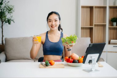 Formda bir Asyalı kadın dijital tabletle çevrimiçi yayın yaparken taze salata, meyve ve meyve suyu içiyor. Parlak, modern bir ev ortamında sağlığın, beslenmenin ve canlılığın vücut bulmuş hali. Sağlıklı bir yaşam tarzı!