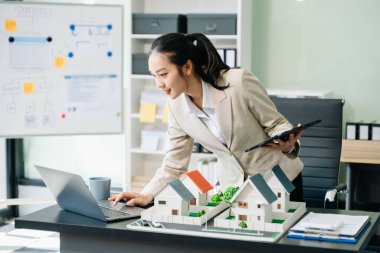 Ofiste dizüstü bilgisayar ve tabletle çalışan genç bir emlakçı ve yanında da küçük ev modelleri..