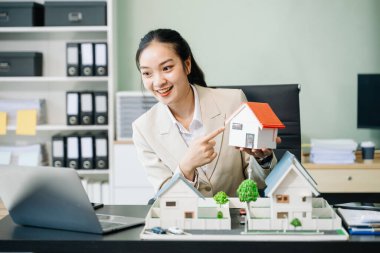 Ofiste dizüstü bilgisayarla çalışan genç bir emlakçı ve yanında da küçük ev modelleri..