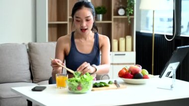 Formda bir Asyalı kadın evde dijital tablet kullanırken taze salata, meyve ve meyve suyu hazırlıyor. Modern ev ortamında sağlık, beslenme ve canlılığın vücut bulmuş hali.