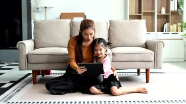 Neşeli anne ve kızı kanepede birlikte vakit geçiriyorlar, gülümsüyorlar ve dijital tablet kullanıyorlar. Aile teması için mükemmel.