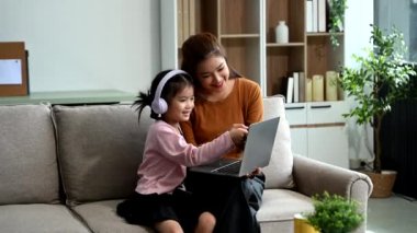 Neşeli anne ve kız kanepede güzel vakit geçiriyorlar, gülümsüyorlar ve kulaklık ve dizüstü bilgisayar kullanıyorlar. Aile teması için mükemmel.